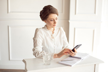 Portrait of a beautiful smiling young brunette businesswoman in a white shirt sitting on a bright modern workstation working with papers .