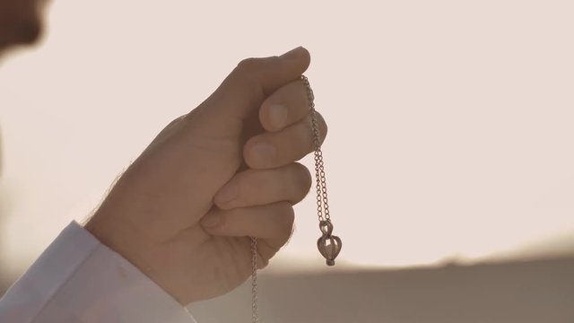 A man looks at a chain with a pendant, inside of which is a pearl. The chain is in his hand, squeezed into a fist. Pendant swaying in the wind. Warm tones. Close-up.