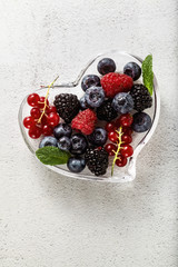 Fresh ripe berries in a glass heart-shaped bowl on a stone white table. summer season. healthy recipes and drinks