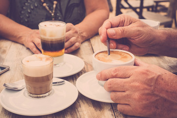 Fototapeta na wymiar Close-up of hands with coffee cups in a cafe bar enjoying cappuccino for tasty breakfast in the morning - aged hands and mature couple have a break together with drinks on a rustic wooden table