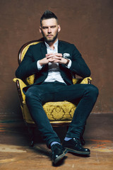 Young handsome man posing for a fashion shoot in a studio. Fashion as a lifestyle. Man wearing a beard. Model sitting on a sofa in tuxedo. Successful fashionable businessman. Business look advertising