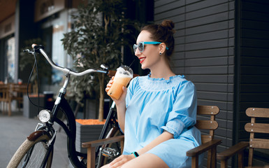 Fototapeta na wymiar Young beautiful lady drinking fresh smoothies on a sunny summer day during her ride. Riding a bicycle in the city. Bicycle tourism. Refreshing fruit drink. Active rest. Hipster on a bike. 