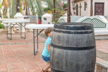 A nice little boy dressed in blue plays hide behind a barrel