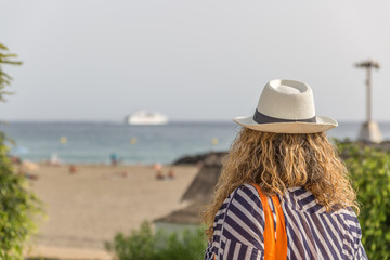 A tourist woman with a hat, placidly observes a beach with people