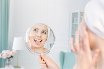 beautiful woman in white bathrobe and towel looking at her perfect skin in mirror , shows the tongue and Applying Tonal Cream Base On Face