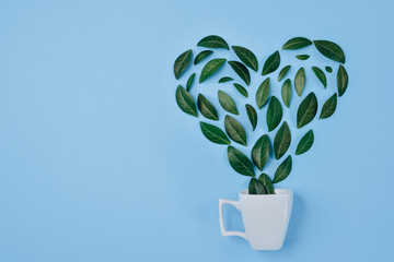 Coffee cup with Green leaves arranged in heart shape over blue background. Love and morning concept. Flat lay, top view, copy space