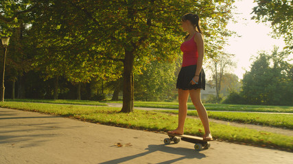 Young woman in a skirt and flip flops riding a skateboard through the park.