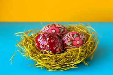 Three red decorated Easter eggs lie in a nest of hay on a yellow and blue background. Ukrainian Easter eggs with ornaments and patterns.