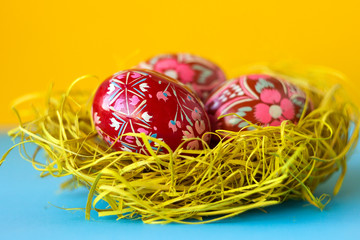 Fototapeta na wymiar Three red decorated Easter eggs lie in a nest of hay on a yellow and blue background. Ukrainian Easter eggs with ornaments and patterns.