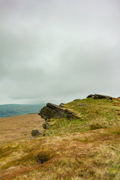 A view of a rocky summit hill with heather and valley in the background under a white cloudy sky