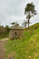 A view of a stone chapel with green vegetation and pine tree under a white cloudy sky