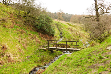 A view of a wooden bridge over a stream with green slopes under a white cloudy sky