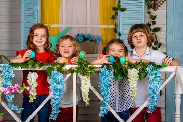 Four beautiful children, two boys and two girls stand on a wooden threshold with Easter eggs in...