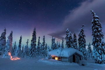 Light shines through window in snow covered cabin under colorful night sky