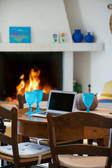 Laptop standing on ancient, vintage and old oak table in cozy living room next to fireplace with glowing, soft burning fire. Empty glasses of wine or beer. Waiting guests. Concept of working at home
