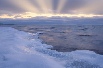 ice and snow on a beach in the foreground, sun rays shining through the clouds in the background