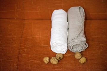 Fototapeta na wymiar Macro shot of rolled, folded snow white and grey natural linen sauna and bath towels next to small walnuts on orange background, on massage table. Beautiful home decor, gift for newlyweds for wedding 