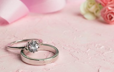 A pair of silver wedding rings on a pink textured surface with flowers and lace decorations in the background.