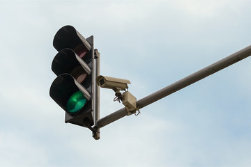 Outdoor surveillance camera and green traffic light both installed on a pole above a roadway, against a blue sky. Modern automatic traffic control.