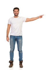 Fototapeta premium Young Man In Jeans And White T-shirt Is Standing, Smiling And Pointing. Front View