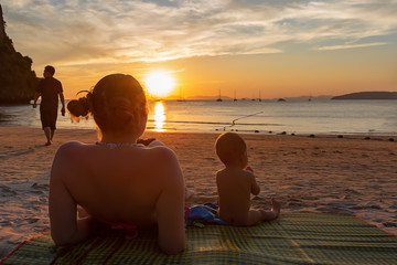 Mother with her daughter. Little baby girl of 9 months. Couple sitting back on a tropical beach, watching and enjoy the sunset. Beautiful blue sky, a bit cloudy, golden sunset light.  Man leaves