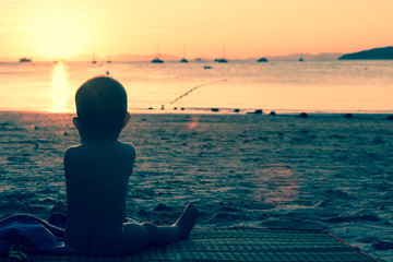 Infant baby girl of nine month old relaxing on the sandy tropical beach. Sitting on the bamboo mat. Sunset and sea, toned in green and red.