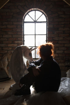 A girl with a unicorn doll sitting in an attic looking out the window