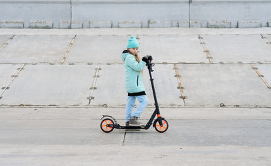 Activities: A school girl with a blue jacket riding a scooter on a gray concrete promenade.