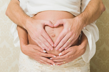 Man holding belly of his pregnant wife making heart. Pregnant woman and loving husband hugging tummy at home. Heart of hands by couple on pregnant belly.