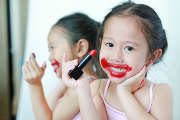 Adorable funny little girl apply lipstick over her mouth with reflection image from mirror.