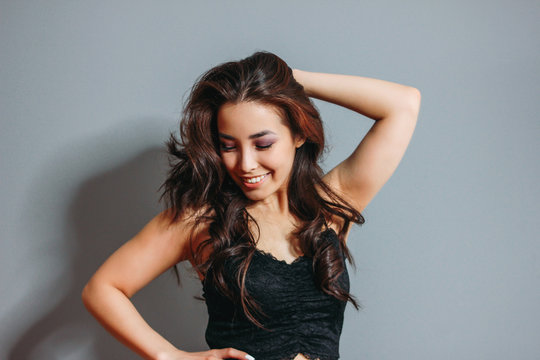 Happy sensual smiling asian young woman with dark long curly hair on grey wall background. Girl having fun at the party