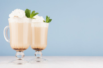Refreshment morning cappuccino coffee with whipped cream and green mint in two exquisite transparent glass in light blue interior, copy space.