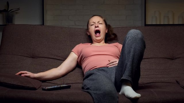 Sleepy woman yawns sitting on the couch watching TV at home. Close up.