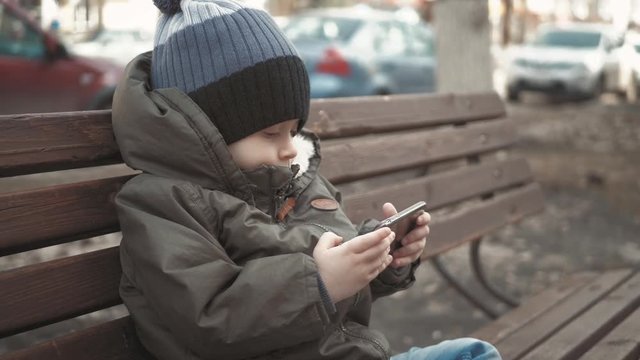 Little boy addictive smartphone sitting on bench in city street. Cute baby boy child with mobile phone on bench outdoor.