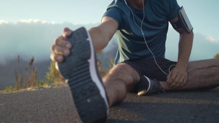 Young happy caucasian brunette man in his athletic clothes preparing to do some exercise outside and stretching his legs whilst sitting down