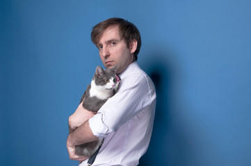 handsome man in pink shirt looking at camera and holding grey cat on blue background with copy space