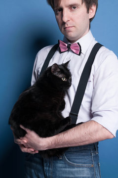 cropped view and side view of man in shirt, suspender and pink bow tie holding cute black cat on blue background 