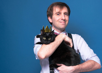 handsome man in shirt and suspenders smiling, holding black cat in golden crown, standing on blue background with copy space