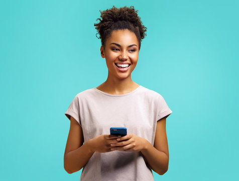 Smiling girl using phone. Photo of african american girl wears casual outfit on turquoise background. Emotions and pleasant feelings concept.