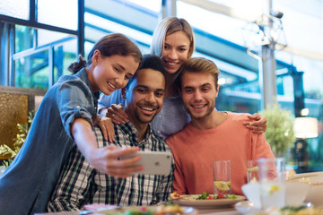 Portrait of four cheerful young friends taking selfie together on smartphone during dinner in cafe