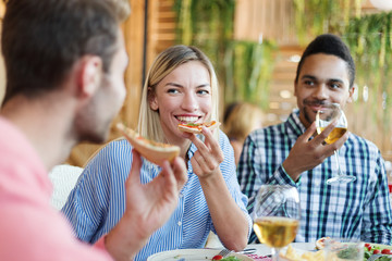 Cheerful young friends enjoying eating pizza and drinking wine during dinner in Italian restaurant