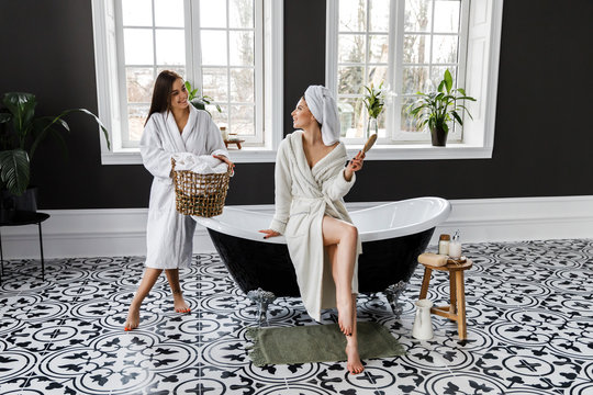 Cute young two women dressed in white dressing gowns and towels on the head have fun in the bathroom