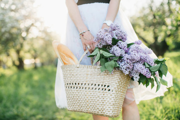 Basket with bouquet of lilacs and baguette in woman hands on background of nature