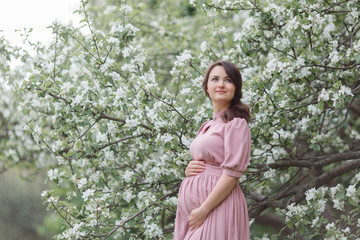 A pregnant woman in a pink dress in an apple orchard is holding a tummy and an apple tree branch in her hands