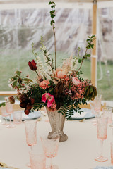 Burgundy and pink flowers centerpiece, pink glasses and blue plates on white table linen. Elegant wedding event decor in marquee, outdoor.