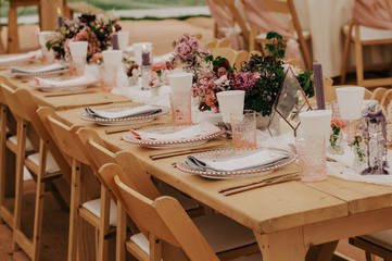 Wedding table settings with burgundy and pink flowers centerpiece, pink glass dinnerware set on wooden table with wooden chairs. Rustic wedding event decor in marquee, outdoor tent.