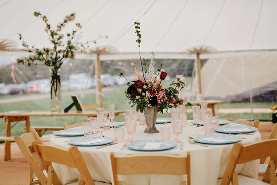 Round banquet table outdoor in marquee tent. FIower center piece, wooden chairs, white linen, simple elegant design for wedding, christenning, birthday celebration. Catering concept