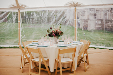 Round banquet table outdoor in marquee tent. FIower center piece, wooden chairs, white linen, simple elegant design for wedding, christenning, birthday celebration. Catering concept