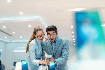 Obraz premium Amazed attractive multicultural couple dressed elegant choosing new smart phone while standing in tech store.
