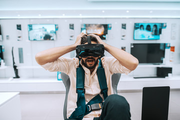 Attractive mixed race bearded man trying virtual reality technology while sitting in chair in tech store.